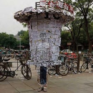 Hiding in the city, info Port 2011 by Liu Bolin, person painted to match the background infront of a message column and bikes