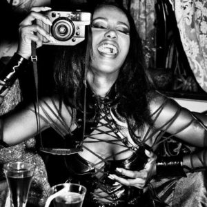 Iman, Paris 1990 by Roxanne Lowit, the model in a black leather bondage top sitting at a table with drinks, holding a camera, laughing