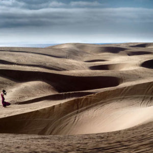 model in red gown running through sand and dunes, the ocean in the back