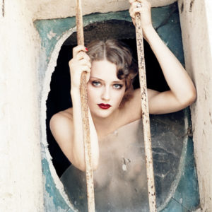 Fräulein by Ellen von Unwerth, nude model looking through a broken oval window, clinging to the windowgrills outside
