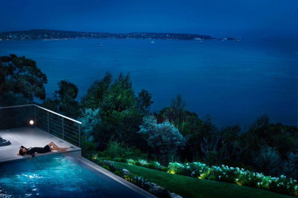 Dreaming of you by David Drebin, model in black dress lying next to a pool in the light, a garden and the ocean in the background