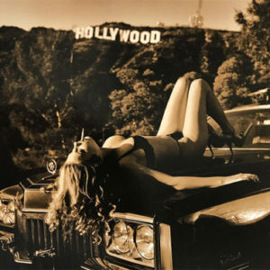 Hollywood Cadillac by Guido Argentini, model in black underwear and heels lying on an engine hood in front of the hollywood sign, smoking