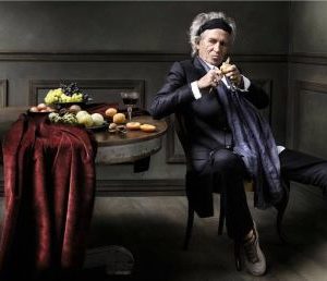 Keith Richards II by Mark Seliger, the singer in a black suit leanig on a table with fruits and red cloth