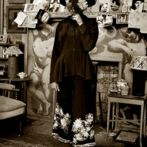 Charlotte - Charleston Farmhouse - England 1987 woman in black embroudered skirt, blouse and hat, holding a bouquet in front of her face, standing in front of a fireplace covered in sketches
