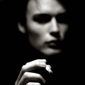 Matthew - Las Vegas 1998 by Albert Watson, blurry black and white portrait of male in high contrast lighting, only his hand holding a cigarette in focus