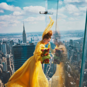 Hadley Robinson IV by Guy Aroch, the actress in a yellow gown, green gloves and floral bag, looking through the glass reiling of a skyscrapers rooftop terrace, the city in the background