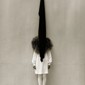Leslie Weiner in Yohji by Albert Watson, girl in white dress and tight with black tulle ruff standing in front of a grey wall with a black piece of fabric hanging over her face