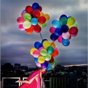Swept away by David Drebin, model in pink dress, tied to coloful air balloones on a balcony over cityscape