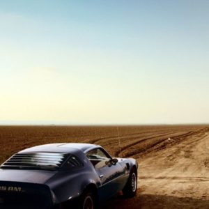 Trans Am by David Drebin, model in short black dress running over an empty field next to a black trans am car