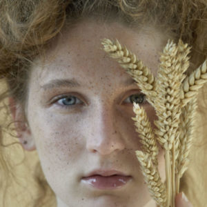 What is Beauty I by Iris Brosch, closeup portrait of model with freckels and messy updo, holding wheat plants in front of her face