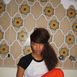 Erykah Badu by Marx Baptiste, portrait of the singer with updo wearing red pants sitting in a bathtub in front of 70s tiles