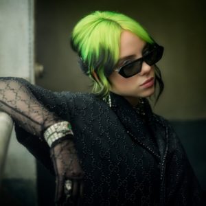 Billie Eilish by Mark Seliger, portrait of the singer with green and black hair, wearing sunglasses and a black gucci shirt with matching gloves