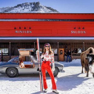 Code Red, Minturn Colorado, 2024 by David Yarrow, Model in red pants with ski standing infront of an oldtimer car, a buffalo and a red Pub