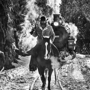 Red Dead 2023 by David Yarrow, Cowboy with gun riding on train trecks infron of an old lokomotive